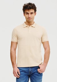Beżowa koszulka polo z kołnierzykiem, krótkimi rękawami i subtelnym logo na piersi. W zestawie z niebieskimi dżinsami.