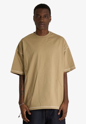 Giovane uomo di colore con capelli corti che indossa una T-shirt beige oversize e pantaloni scuri, in piedi su uno sfondo chiaro uniforme.