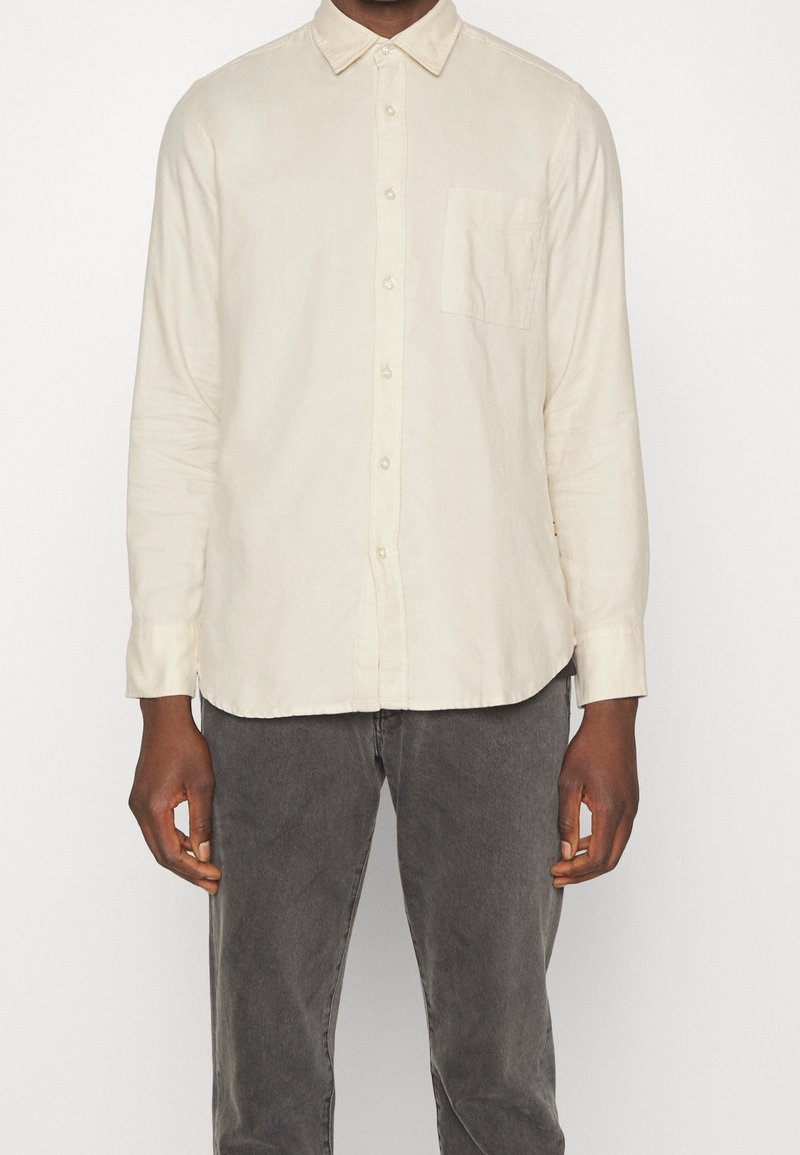 Homme portant une chemise crème à manches longues boutonnée et un pantalon gris foncé, debout devant un fond blanc uni.
