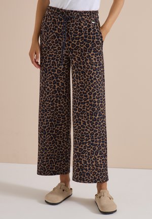 Frau trägt braune und schwarze Leopardenmuster-Hose mit weitem Bein, Kordelzug in der Taille und beige Slipper auf einfarbigem Hintergrund.