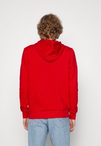 Sudadera roja con capucha ajustable, puños de canalé y dobladillo. Tejido suave con una textura lisa, llevada sobre unos vaqueros azul claro.