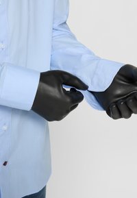 Lyseblå skjorte med lange ermer og knappelukking, glatt tekstur og franske mansjetter. Svarte hansker justerer mansjetten.