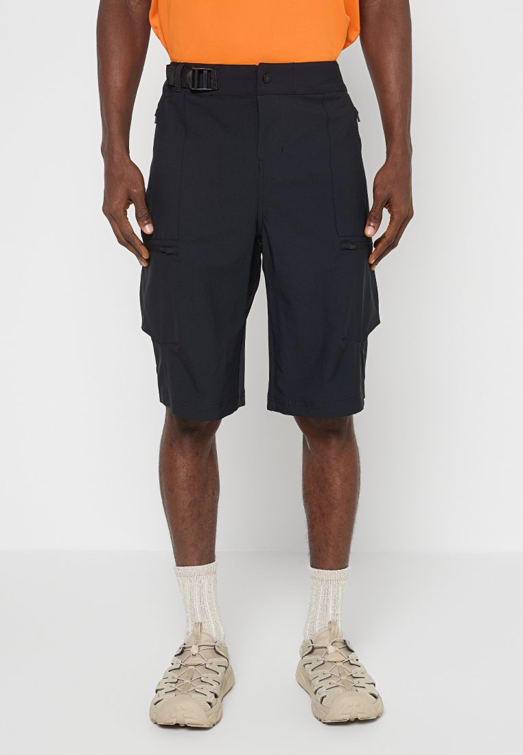 Homme portant un short cargo noir avec des poches latérales, des chaussures de randonnée beiges, des chaussettes blanches et une chemise orange.