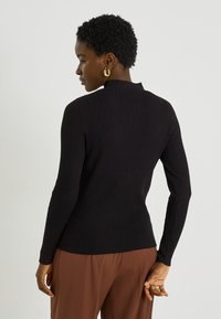 Top negro de manga larga con costuras acanaladas y cuello alto, con un diseño ajustado y tejido texturizado. La modelo lleva pantalones marrones.