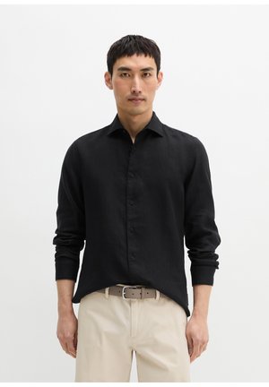 Mężczyzna stoi, ubrany w czarną, długą koszulę zapinaną na guziki, beżowe spodnie i brązowy pasek na jednolitym białym tle.