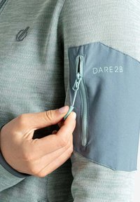 Grau gefütterte Armtasche auf einem strukturierten grünen Pullover, mit einem Logo "DARE2B" in hellgrau. Tasche mit einem türkisfarbenen Zugband akzentuiert.