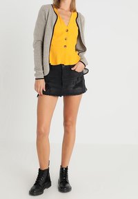 Cardigan beige texturé avec bordures noires, haut jaune à boutons, short en denim noir effiloché et bottines noires à lacets.