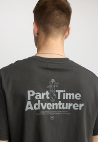 Camiseta gris gráfica de manga corta, con el texto "Aventurero a Tiempo Parcial" y un personaje de dibujos animados impreso en la parte posterior en blanco.