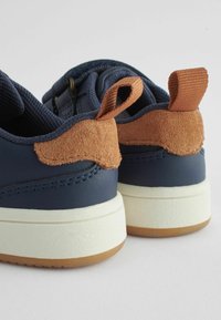 Baskets bleu marine avec un accent en daim beige à l'arrière, semelle en caoutchouc blanc et languette en mesh texturé. Comprend des tirettes pour un enfilage facile.