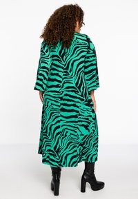 Lange jurk met groen en zwart zebra-print en losse mouwen, met een vloeiende silhouette en kniehoge zwarte platformlaarzen.
