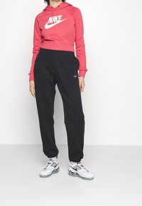 Červená krátká mikina s kapucí a bílým logem Nike, doplněná černými tepláky s bočními kapsami a malým bílým logem Nike na noze.