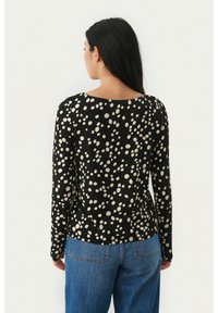 Woman with long dark hair wearing a black long-sleeve top with white irregular dots and blue jeans, facing away against a plain background.