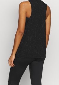 Canotta sportiva nera con vestibilità ampia, design senza maniche e tessuto testurizzato. Presenta una schiena aperta per la ventilazione. Abbinata a leggings neri.