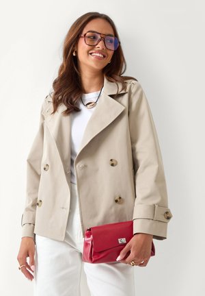 Mujer sonriendo, vistiendo una chaqueta beige cruzada, camisa blanca y pantalones, gafas grandes y sosteniendo un bolso rojo.