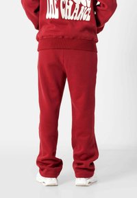 Person steht in roten Jogginghosen und passendem Sweatshirt mit weißem Schriftzug, kombiniert mit weißen Sneakern, vor einem schlichten Hintergrund.