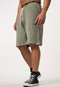 Shorts de toalla en color verde oliva con una cintura elástica negra, cordón blanco y detalle de bolsillos laterales. Llevados con zapatillas altas negras.