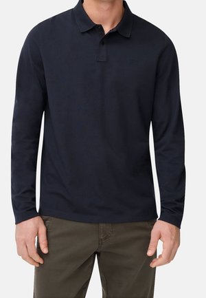 Homme portant un polo bleu marine à manches longues avec boutons, associé à un pantalon vert olive foncé, debout devant un fond blanc.