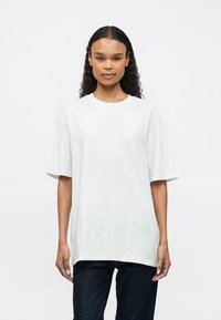 Žena s dlhými kučeravými vlasmi, oblečená v oversize bielom tričku a tmavých džínsoch, stojí na pozadí jednoduchého bieleho pozadia.