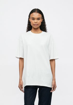 Žena s dlhými kučeravými vlasmi, oblečená v oversize bielom tričku a tmavých džínsoch, stojí na pozadí jednoduchého bieleho pozadia.