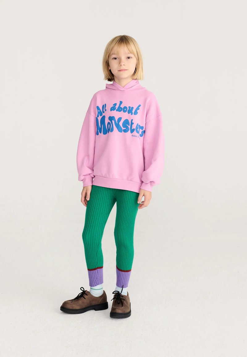 Enfant portant un sweat à capuche rose avec le texte "All about Monster", des leggings verts côtelés avec des poignets violets, des chaussettes blanches et des chaussures marron, debout sur fond blanc.