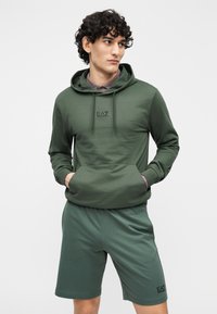 Ciemnozielona bluza z kapturem z kieszenią typu kangur i troczkami, z logo EA7. W zestawie pasujące ciemnozielone spodenki, gładka faktura tkaniny.