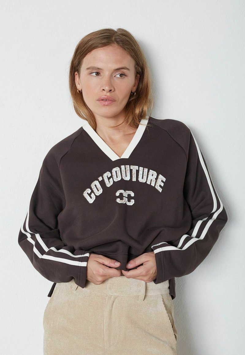 Mujer que lleva un sudadera oscura con cuello en V y rayas blancas en las mangas, con el texto "CO'COUTURE", combinada con pantalones de pana beige.
