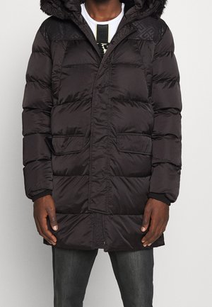 Doudoune noire longue d'hiver avec poches avant portée sur une chemise blanche et un jean foncé, visible du cou jusqu'à mi-cuisse.