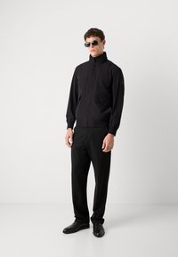 Juodas zip-up džemperis su aukštu apykakle, priekinėmis kišenėmis ir elastinėmis rankovėmis, dėvimas su tamsiomis kelnėmis ir elegantiškais juodais bateliais.