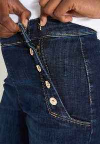 Jean en denim bleu foncé à taille haute, avec une fermeture à boutons, des boutons dorés et des coutures contrastantes jaunes. La texture est lisse.