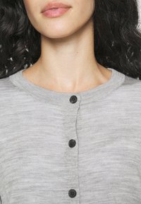 Cardigan gris en tissu texturé, avec un col rond et cinq boutons noirs le long de l'avant. Gros plan sur le col et les boutons.