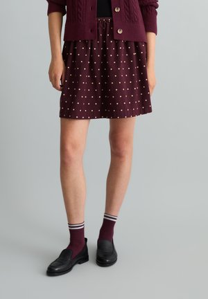 Frau trägt einen burgunderfarbenen Rock mit Punkten, passende Strickjacke, gestreifte Socken und schwarze Loafers, steht vor einem schlichten Hintergrund.