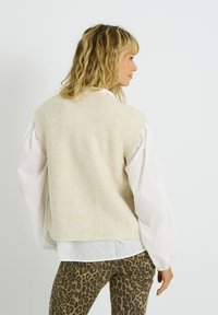 Mujer de perfil hacia la derecha, con un chaleco de punto beige sobre una blusa blanca y pantalones de estampado de leopardo, frente a un fondo blanco liso.
