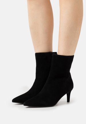 Stivaletti neri in suede con punta a punta e tacchi bassi a kitten indossati su gambe nude su uno sfondo bianco.