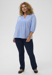 Blouse bleu clair, ample, avec un col en V, des plis et des manches trois-quarts, associée à un jean bleu foncé et des ballerines noires.