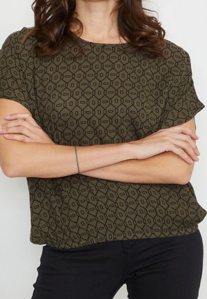 Personne portant une chemise verte à manches courtes à motifs, les bras croisés et un pantalon noir, montrant un fin tatouage bracelet sur un poignet.
