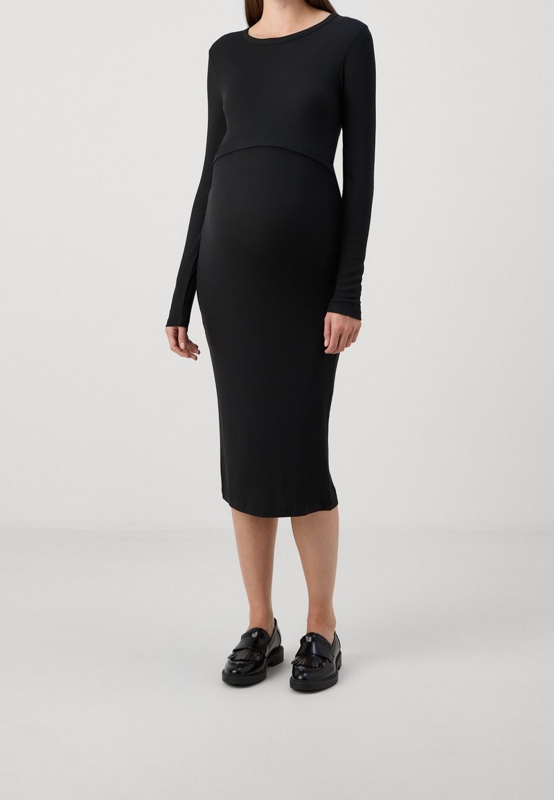Femme portant une robe midi noire ajustée à manches longues et des mocassins noirs, debout devant un fond gris clair uni.