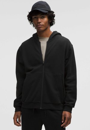 Młody mężczyzna noszący czarną bluzę z zamkiem, jasnoszarą koszulkę, czapkę z daszkiem, stoi na tle jednolitego jasnoszarego tła.