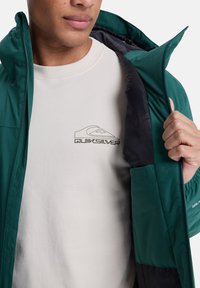 Man in een witte Quiksilver-shirt, met een open donkergroene jas met zwarte voering, gedeeltelijk gezicht en hand zichtbaar.