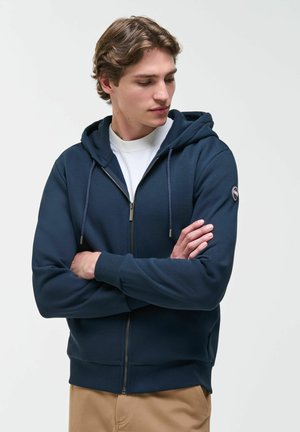 Giovane uomo con capelli castano chiaro che indossa una felpa con cappuccio blu navy con zip e pantaloni beige, braccia incrociate, guarda verso il basso e di lato.