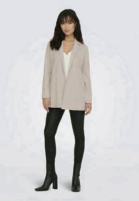 Blazer bege claro com um design elegante, lapelas recortadas e mangas longas, usado sobre uma blusa branca e calças pretas ajustadas, com botas de tornozelo.