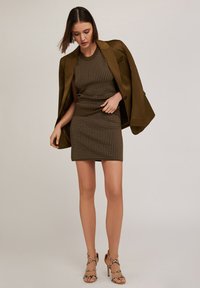 Brun stickad klänning med vita prickar, matchad med en mörk olivgrön blazer. Outfiten har en figurnära silhuett och sandaler med remmar och hög klack.