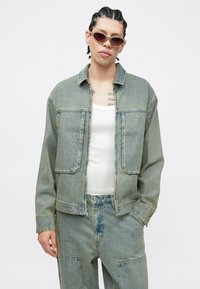 Τζιν μπουφάν και παντελόνι σε ανοιχτό μπλε με ξεθωριασμένες αποχρώσεις. Διαθέτει δύο εμπρόσθιες τσέπες και κλείνει με κουμπιά. Το μοντέλο φοράει γυαλιά ηλίου.