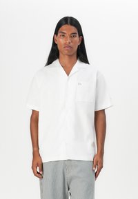 Camicia bianca a maniche corte con bottoni, dotata di tasca e un logo discreto, realizzata in tessuto leggero. Shorts a righe completano l'outfit.