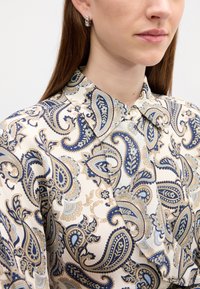 White blouse with a blue and beige paisley pattern, featuring a collar and buttons, paired with silver stud earrings.