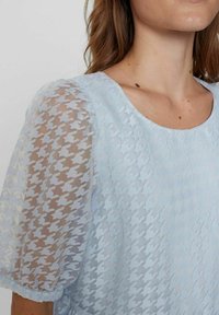 Svijetloplava bluzica od teksturiranog materijala s motivom houndstooth, kratkim puf rukavima od prozirnog materijala i okruglim izrezom.