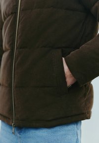 Brun fløjls-pufferjakke med lynlås foran, som har en sidelomme og en blød, tekstureret finish, parret med lyseblå jeans.