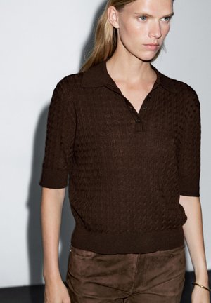 Pull-over polo en tricot brun avec col, manches courtes et motif texturé. Porté avec un pantalon en daim brun assorti.