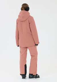Veste de ski rose avec capuche, assortie à un pantalon. Présente des zips latéraux et des accents noirs sur le pantalon et les chaussures. Texture lisse.