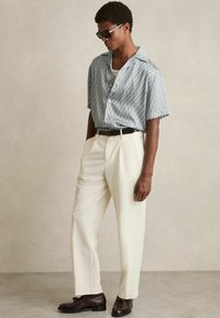 Cămașă cu mânecă scurtă, cu nasturi, în model geometric alb și albastru, asortată cu pantaloni crema și pantofi maro închis, având un curea neagră.