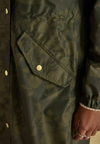 Artimųjų planų tamsiai žalia gėlėmis raštuota paltinė kišenė su auksiniu spalvos užsegimu ir elastingu rankogaliu, kurioje matoma ranka su sidabriniais žiedais.